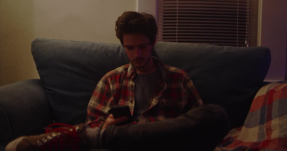 Man sits on a blue couch near a window looking at his phone