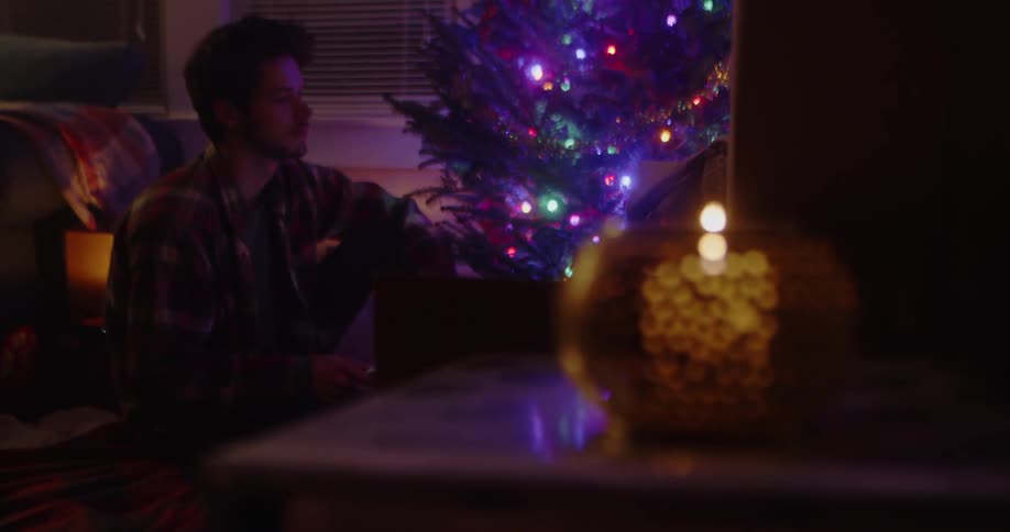 Man sits near a lit up christmas tree and a candle burns on a table