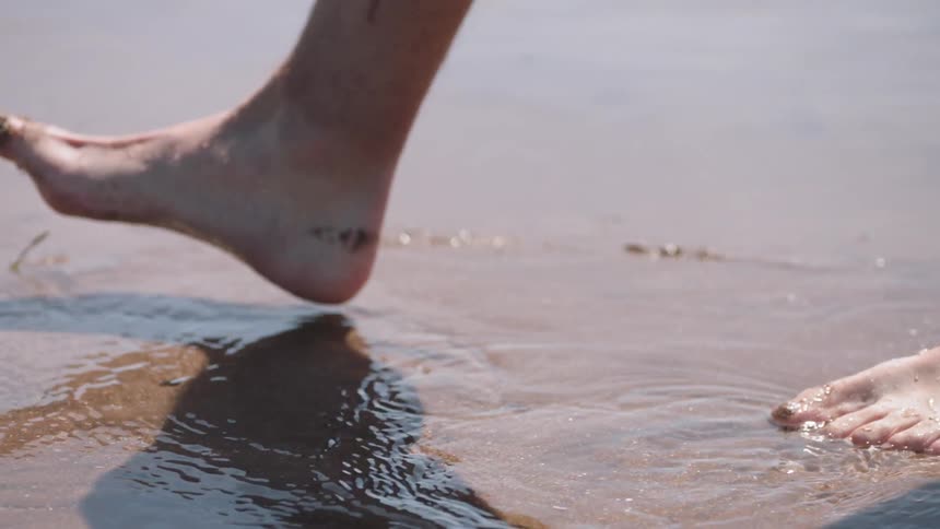 Person's feet step into shallow water and sandy mud on a beach