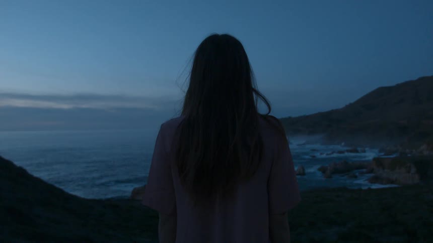 A woman stands on the cliff looking out onto the water
