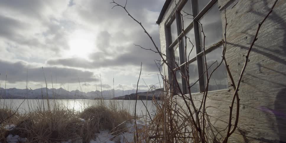 The sun is shining through clouds onto a cabin on a shoreline in winter