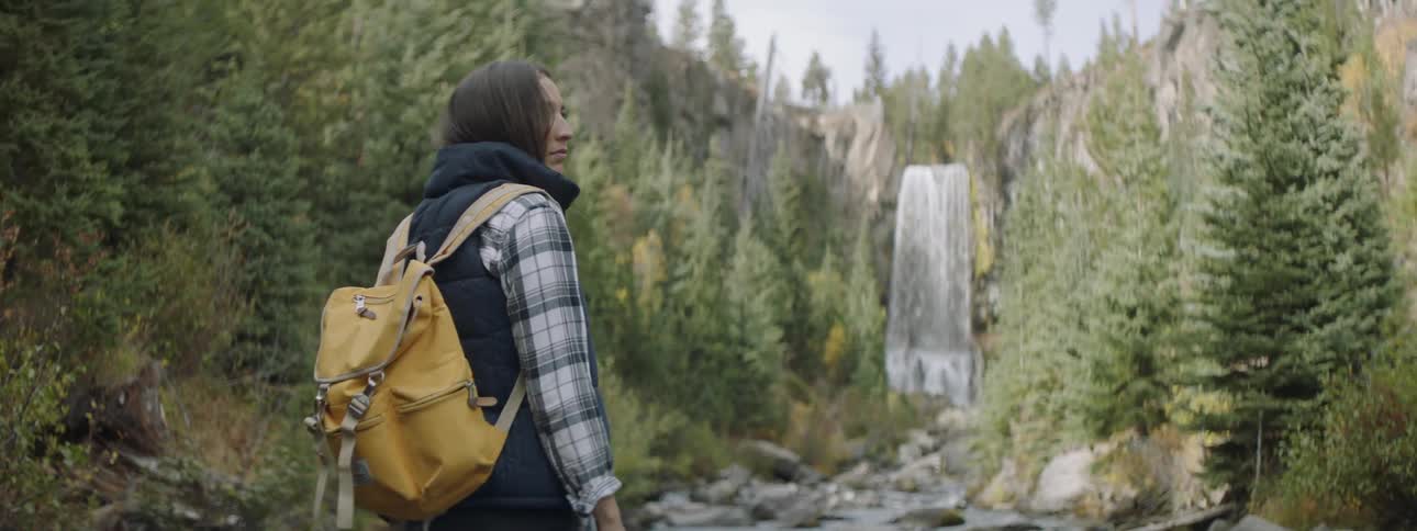 Woman wearing a yellow backpack stands in the woods near a waterfall