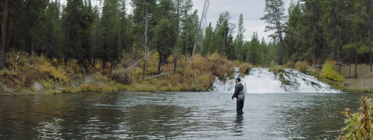 Man is fly fishing in a river near a small waterfall