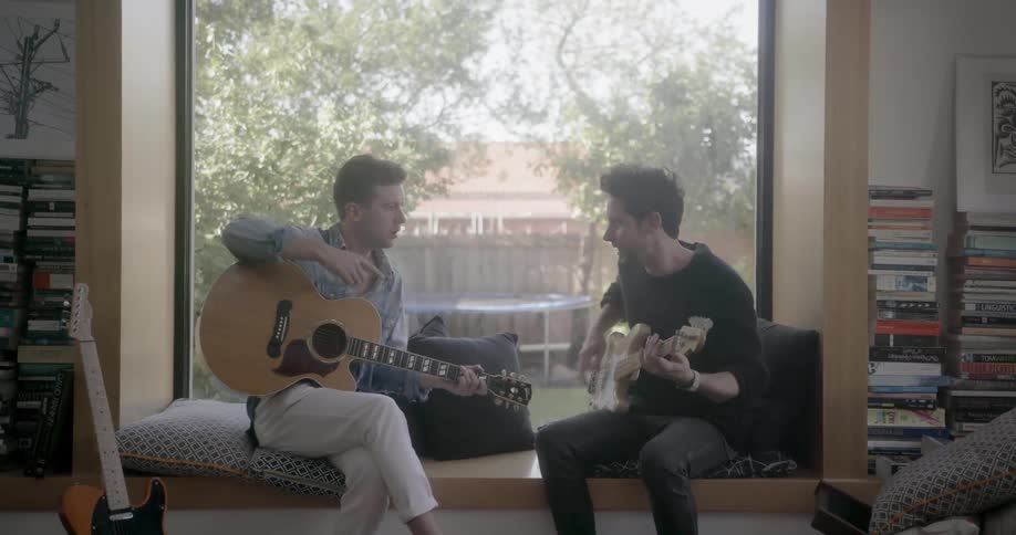 Two men play guitar together while sitting on a window seat