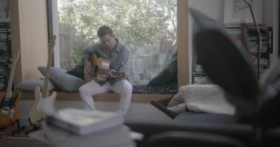 A man sings and plays guitar sitting in a window seat