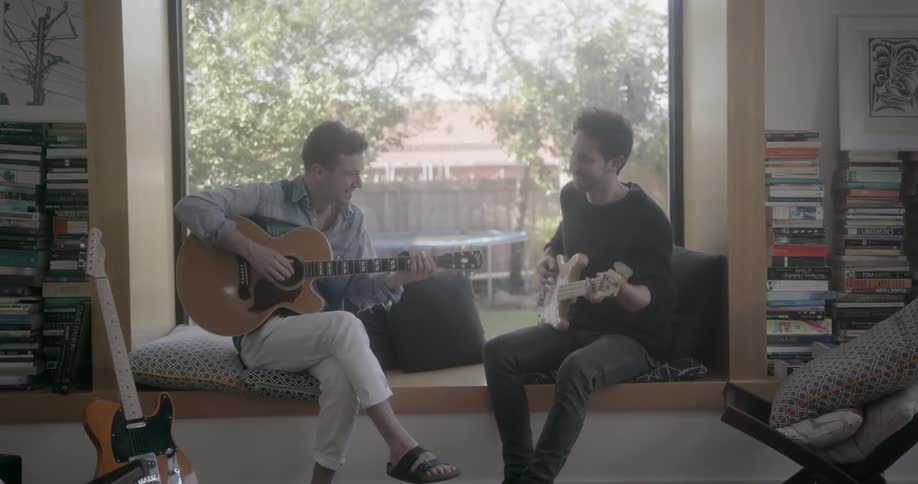 Two men sitting on a window seat play guitar and talk