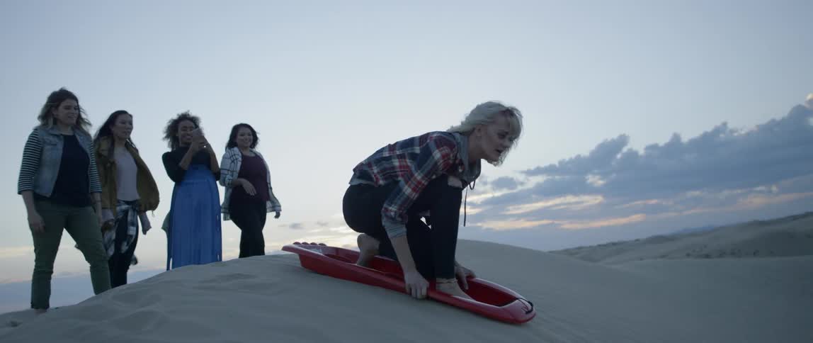 Woman tries to push a sled down a sandy hill with a group of women behind her watching