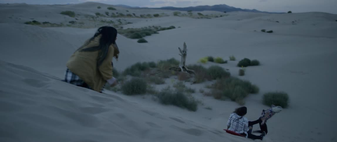 Women sliding down a sandy hill on a sled as other watch