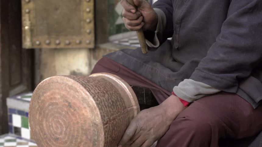 An artisan is hammering a pattern into a copper pot during the day