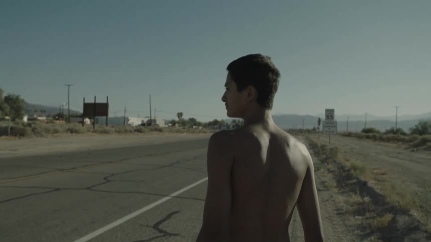 A shirtless teenage boy walks down a road