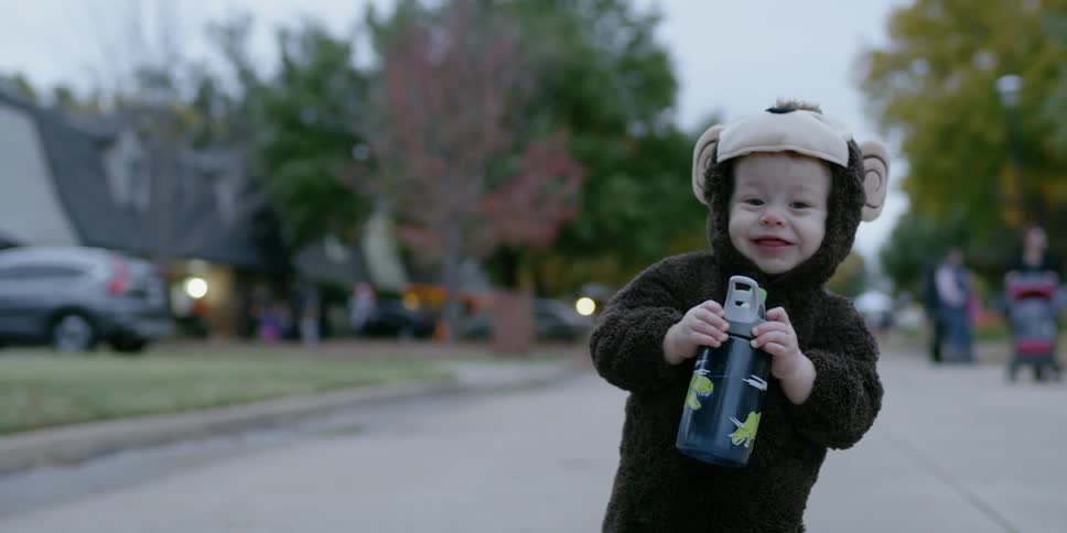 A toddler is sipping from a drink bottle as he stands outside at dusk on halloween
