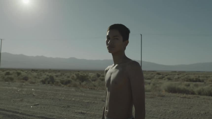 A young man stands shirtless on a dirt road on a sunny day