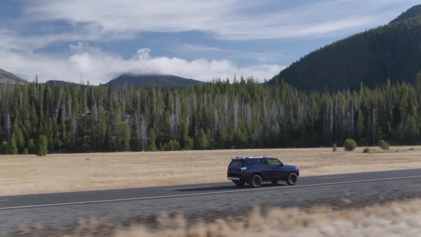 An suv is driving on a road past a forest and cloud covered mountain during the day