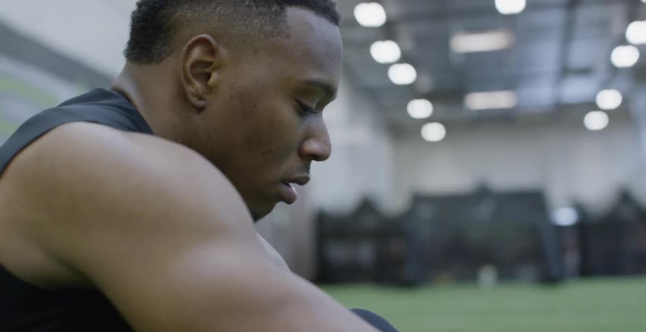 A male athlete is sitting on an indoor football field putting on his shoes