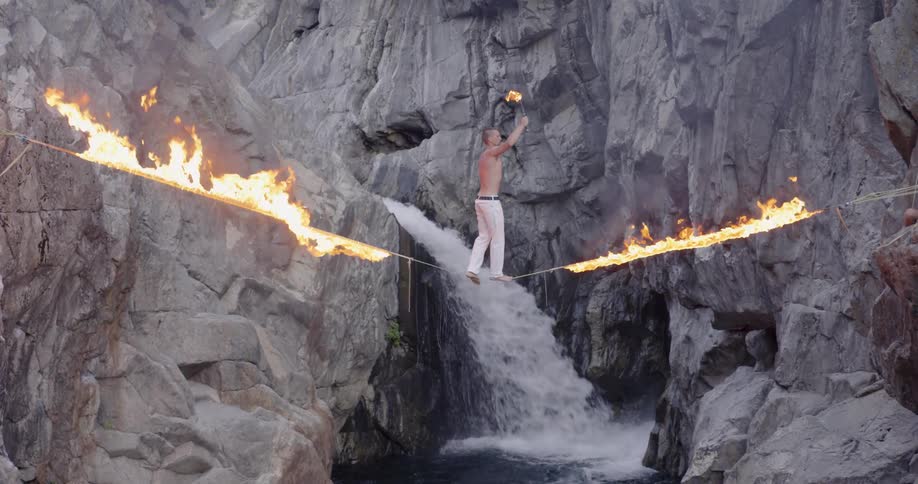 A man is on a burning slack line over a waterfall and breathing fire