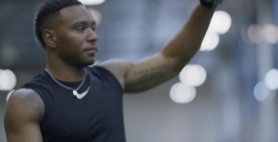 A male athlete is holding a football and stretching his arm indoors during the day