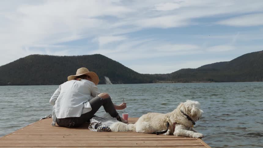 A woman sits on a dock with her dog and paints near the water