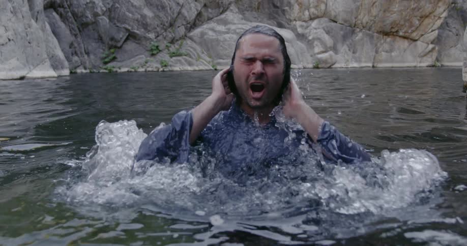 A man is surfacing from a pool of water fully dressed in a natural setting during the day