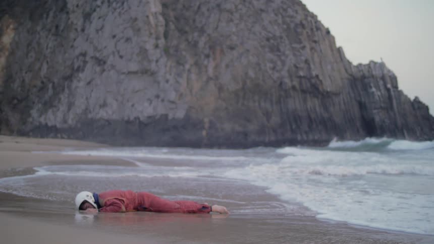 Person wearing helmet is lying face down in the sand as waves hit