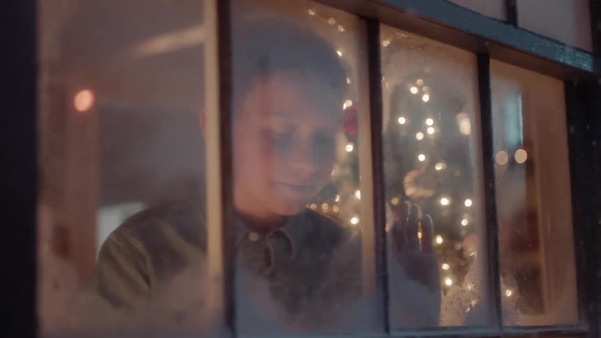 A boy looks out a window and watches it snow on christmas