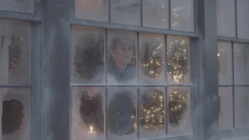 A boy looks out a window while it snows on christmas