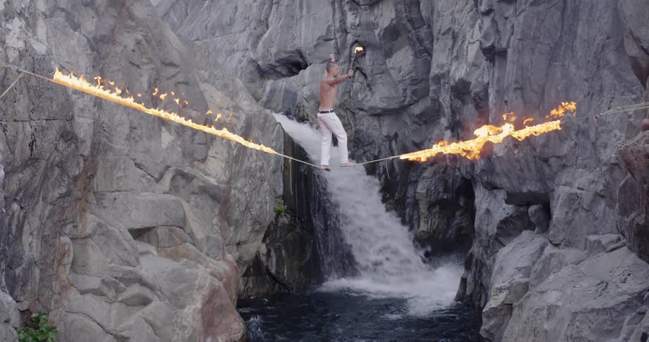 A man is balancing on a burning tightrope over a gorge with a waterfall
