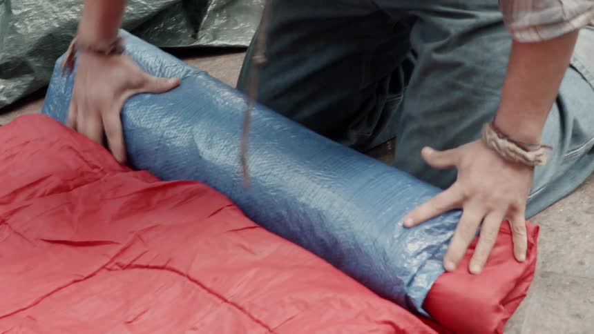A man kneels on the ground and rolls up a sleeping bag