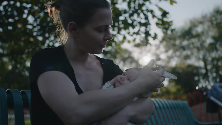 A woman holds a newborn on a park bench at sunset