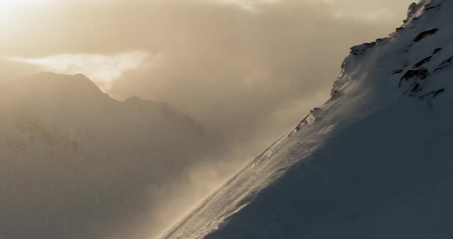 Snow blows past a mountain ridge at sunset