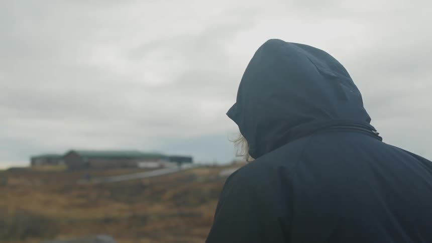 A person in a hood and coat walks across a countryside towards a building in the distance on a cloudy day