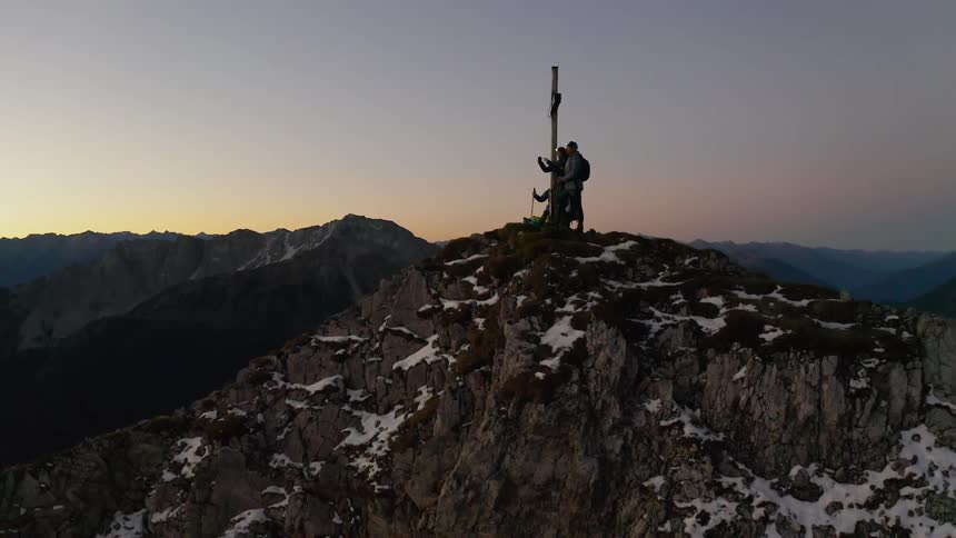 Hikers stand next to a cross on a mountain peak at sunset
