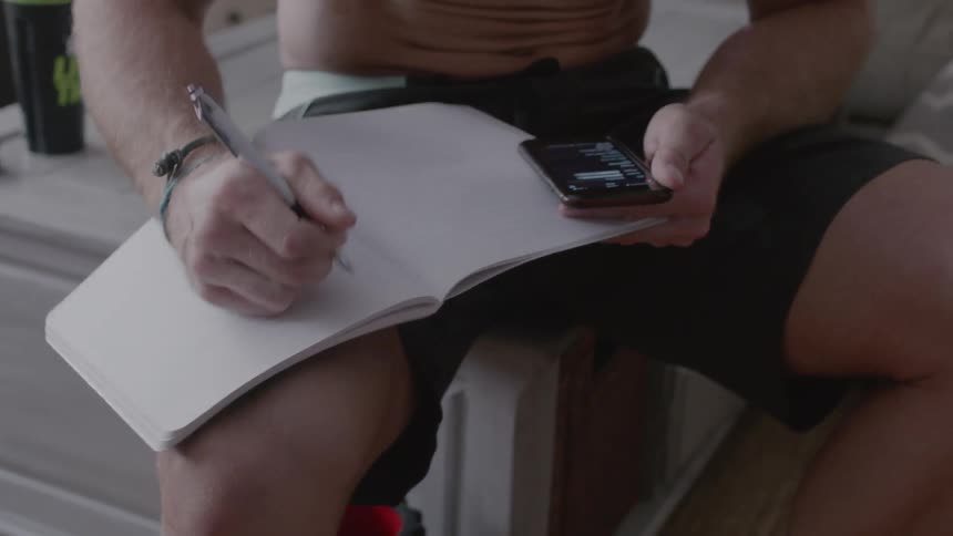 A man is holding a phone and writing in a notebook during the day