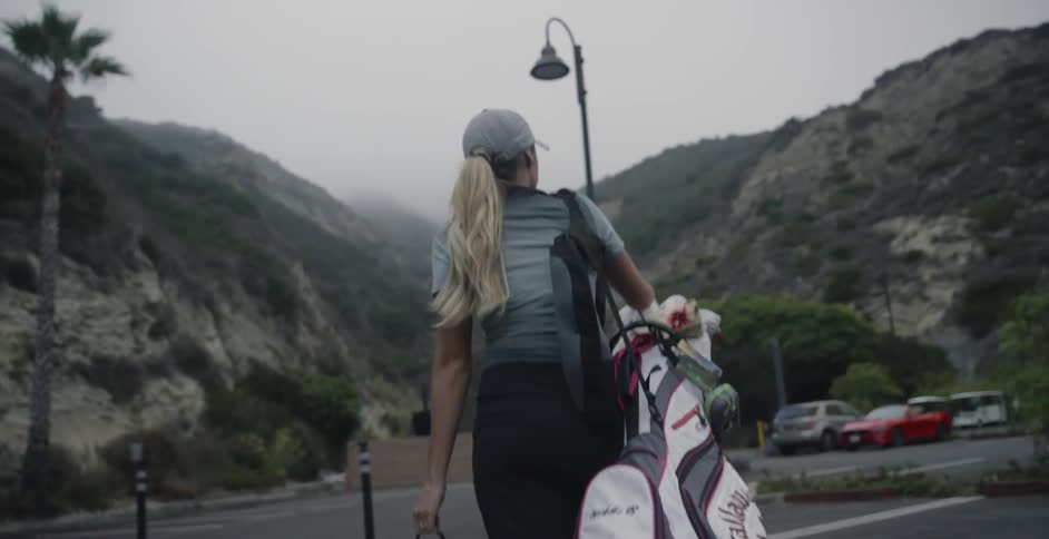 A woman walks in a parking lot with golf clubs and a bucket of golf balls