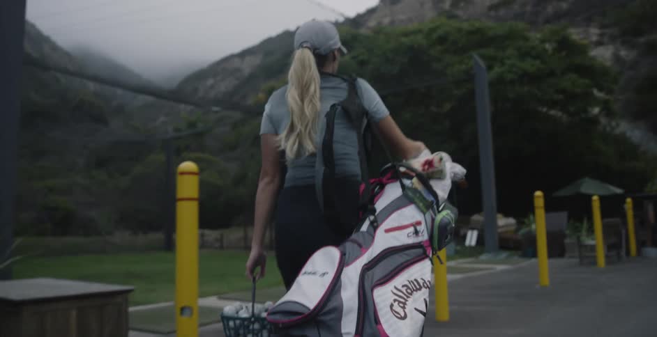 A female golfer is carrying a golf bag and bucket of balls towards a driving range