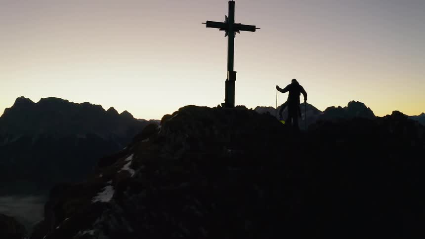 Hikers walk next to a cross on a mountain peak at sunset