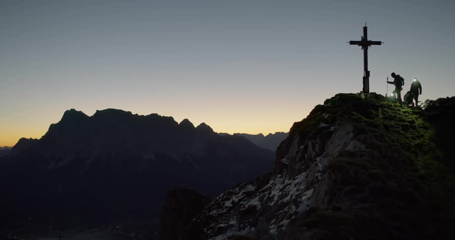 Hikers climb up to a cross on a mountain peak at sunset