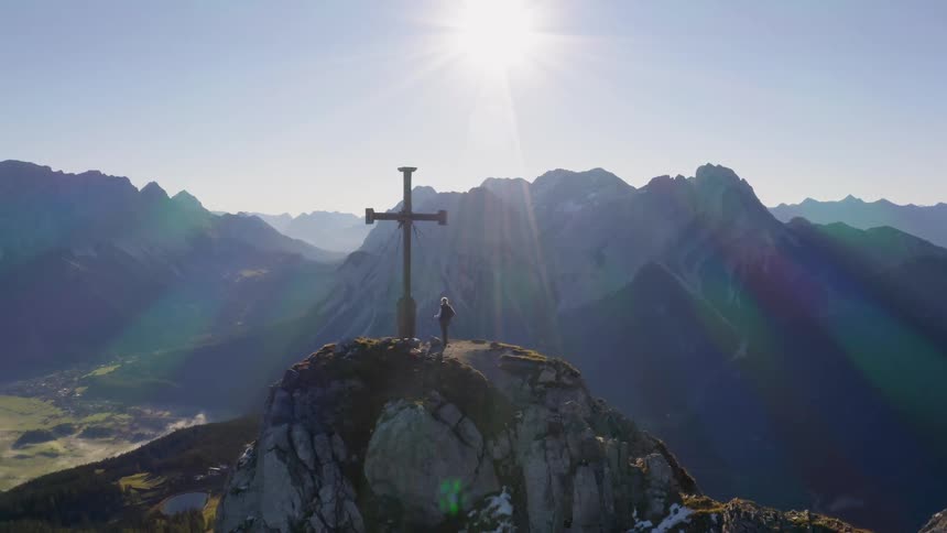 A person is standing near a cross on a mountain peak in daylight