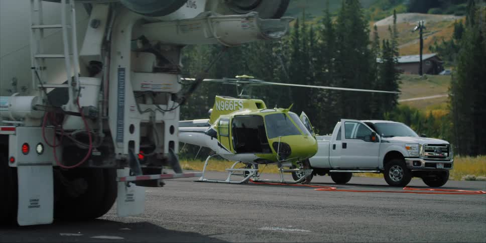 A cement truck is rolling backwards on a tarmac past a helicopter during the day