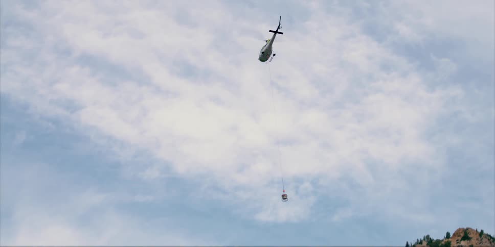 A helicopter with a basket hanging from a cable flies over trees and ridge