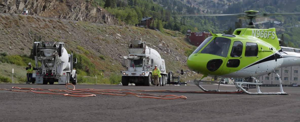 Helicopter spinning its propeller and several cement trucks in a mountainous area