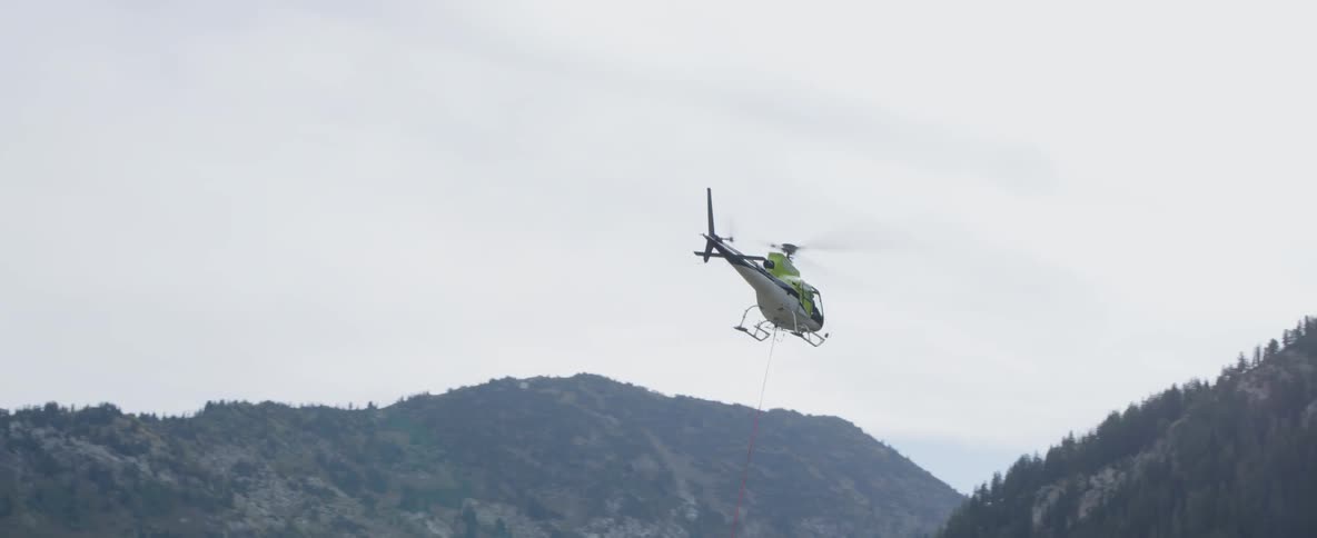 A helicopter takes off in the mountains with a long red cable hanging below it
