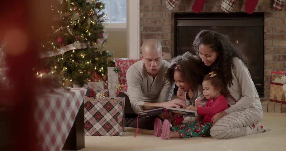 A family are reading a story together next to a christmas tree and presents