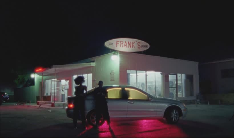 Two people are leaning up against a car outside a laundromat at night