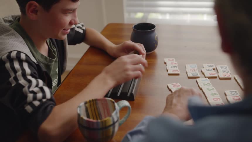 A man and a boy sit at a table playing a game with numbered tiles