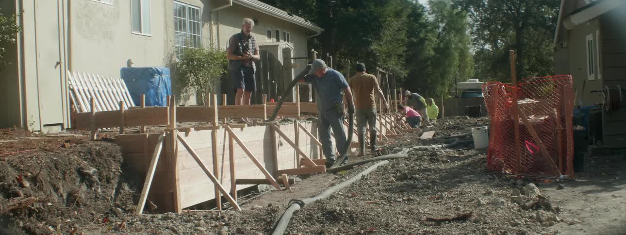 Workers fill a framed wall with cement on a sunny day