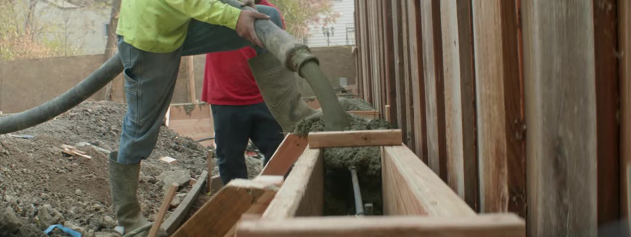 A worker pours cement from a hose as another shovels behind