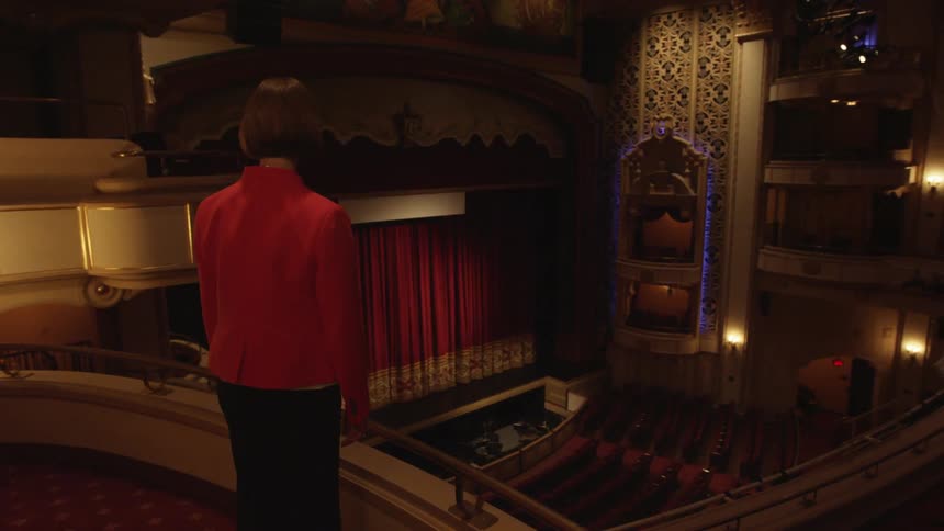 Woman stands on a balcony in a theatre and looks out at the seats and stage below