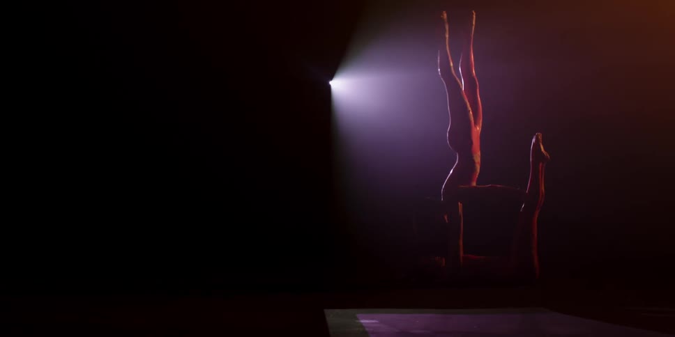 Two women perform acrobatics in a dark room with a light behind them