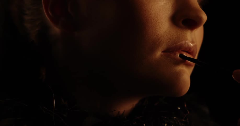 A woman in a dark room has lip gloss applied to her lips slowly