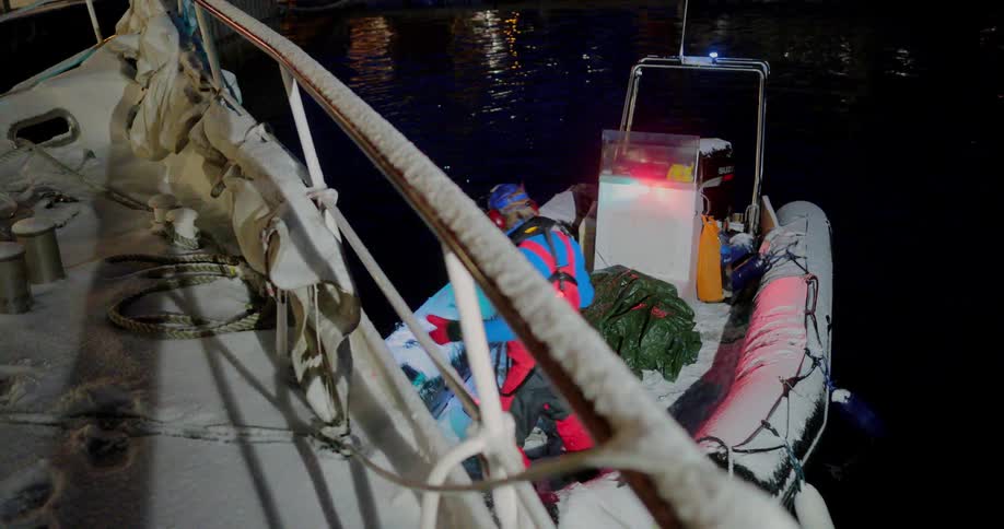 A man on a small boat holds a rope as he brings it close to a larger boat at night